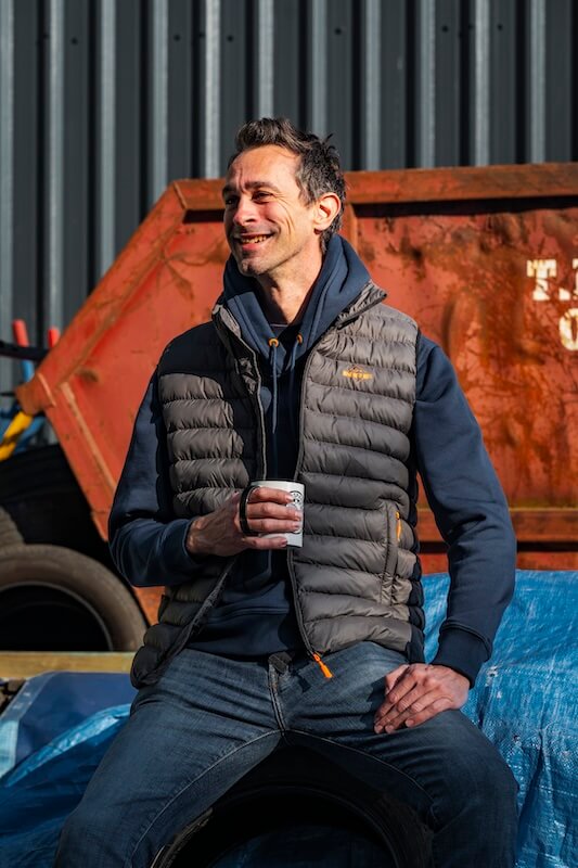 A Man wearing a gillet drinking coffee in front of a skip